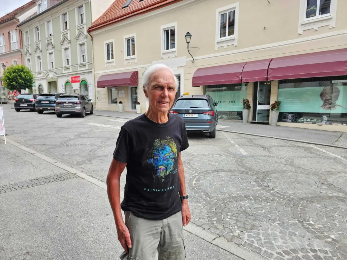 Ein älterer Mann mit weißem Haar steht auf einer Kopfsteinpflasterstraße, trägt ein schwarzes T-Shirt mit einem farbenfrohen Design. Hinter ihm stehen Gebäude mit roten Vordächern an der Straße, und ein geparktes Auto ist zu sehen.
