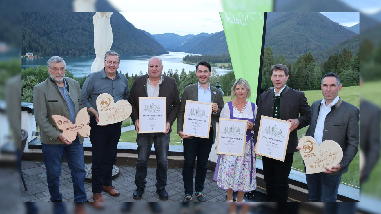 Fünf Personen halten Urkunden vor einer malerischen Berg- und Seekulisse. Die Urkunden haben grüne Ränder und enthalten Logos und Text.