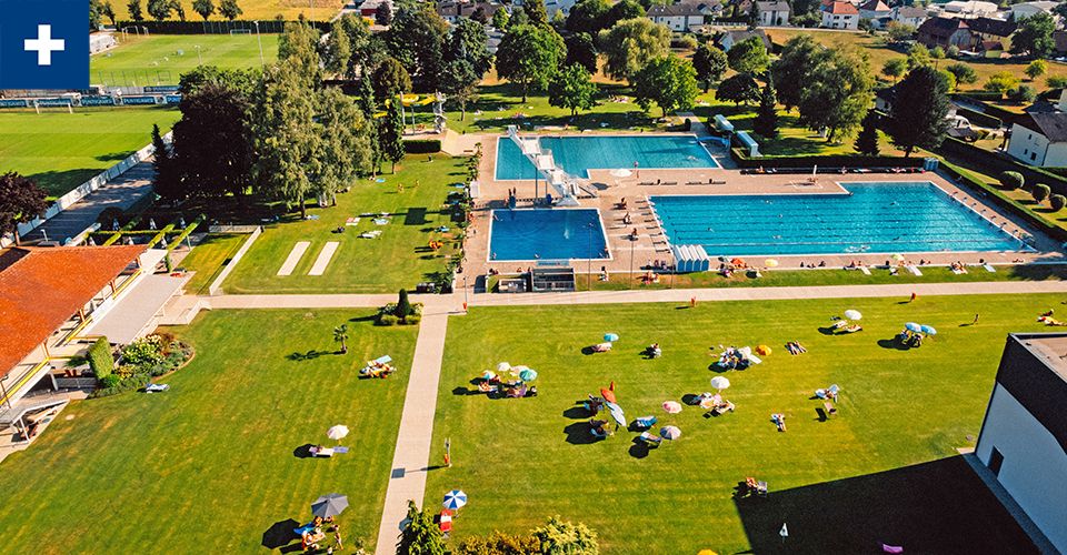 Eine Luftaufnahme eines öffentlichen Schwimmbads mit mehreren Bahnen. Menschen schwimmen, sonnenbaden und sitzen auf der Wiese.