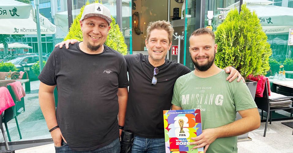 Drei Männer posieren für ein Foto mit einem FIFA-Weltmeisterschaftsbuch 2026, lächelnd und stehen vor einem Gebäude.