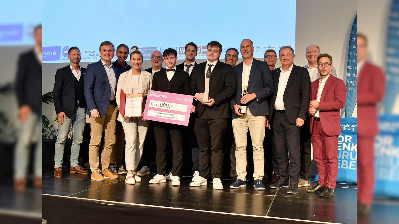 Eine Gruppe von Männern und Frauen in formeller Kleidung steht auf der Bühne und hält Auszeichnungen und ein Zertifikat. Ein Mann hält einen pinkfarbenen Scheck über 1.000 Euro und ein anderer eine Flasche.
