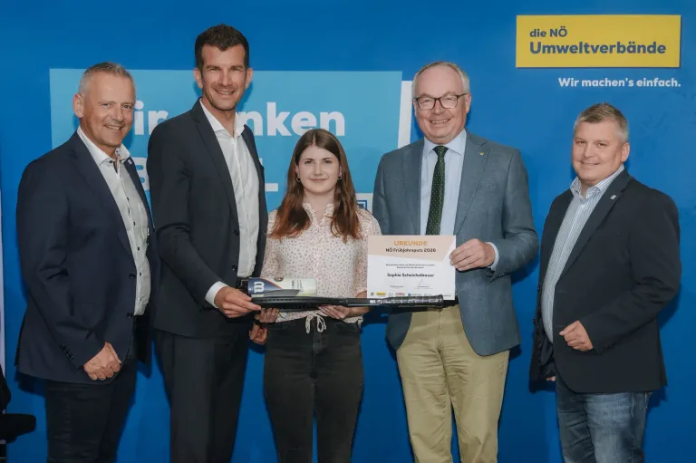Fünf Personen stehen hinter einem blauen Hintergrund, zwei Männer auf beiden Seiten eines Mädchens, das ein Zertifikat und einen Tennisschläger hält. Der Mann auf der linken Seite trägt einen schwarzen Anzug und ein weißes Hemd.