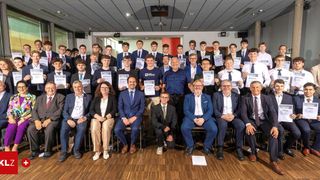 Eine Gruppe von Menschen in formeller Kleidung, die Zertifikate mit gedruckten Jahren in den Händen halten. Einige Leute sitzen auf Stühlen, während andere stehen.