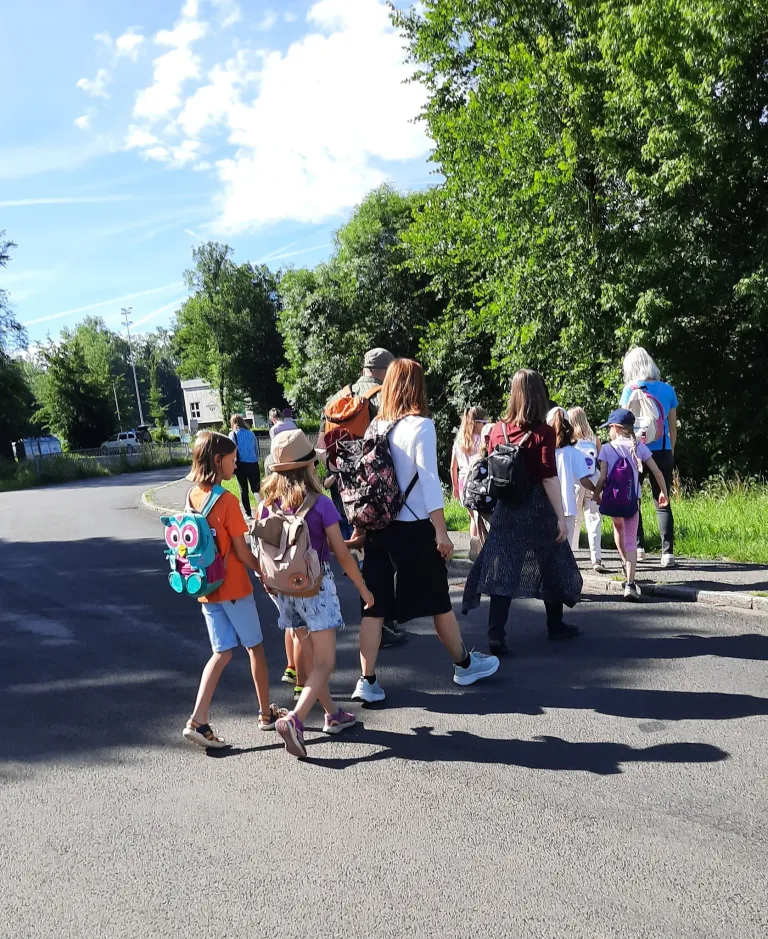 A group of children and adults walk on a road, wearing backpacks, under a blue sky with some clouds. Trees line the sides of the road.