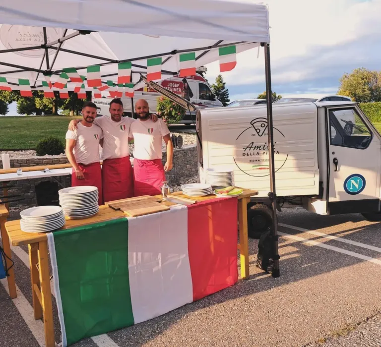 Drei Männer in roten Schürzen stehen hinter einem Tisch mit Tellern und einer Flagge. Ein Pizzawagen ist in der Nähe geparkt.