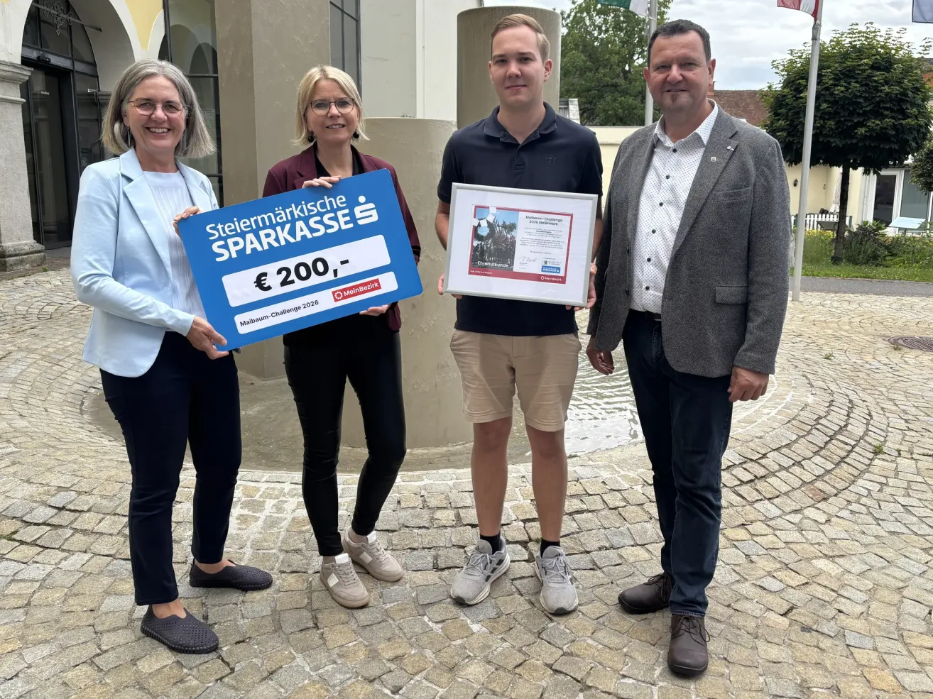 Drei Personen stehen zusammen mit einer Plakette und einem blauen Schild mit weissem Text, der 'Steiermarkische SPARKASSE' und eine Geldsumme von '200 Euro' liest.