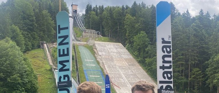 Two people stand with snowboards in front of a ski jump surrounded by trees and a slope.
