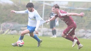 Zwei Fußballspieler sind auf einem Rasenplatz. Einer trägt ein weißes Trikot, der andere ein bordeauxfarbenes. Sie kämpfen um einen Fußball.
