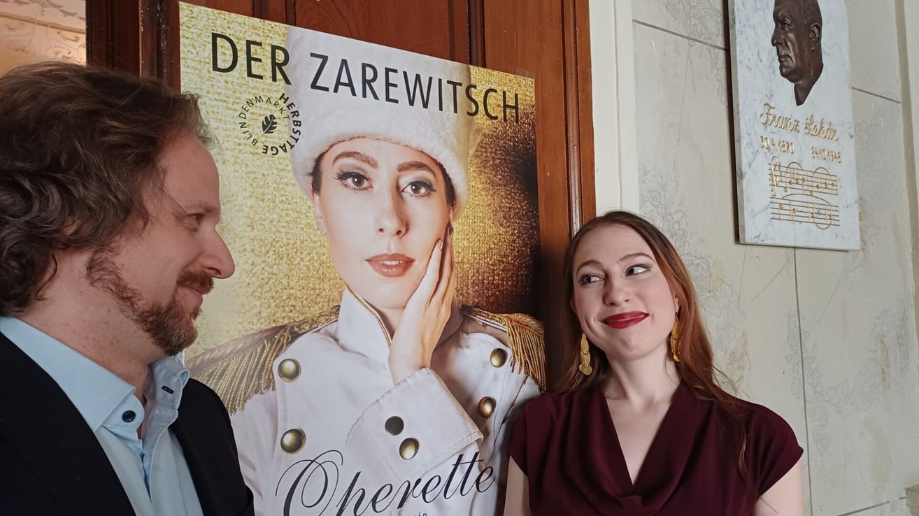 Drei Personen stehen vor einem Plakat für die Oper 'Der Zarewitsch' mit einer Frau in Uniform.