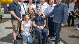 Eine Gruppe von Menschen in Uniformen und mit Brillen steht und sitzt auf dem Bürgersteig, lächelt und posiert für ein Foto. Ein Mann im Rollstuhl ist ebenfalls im Foto enthalten.