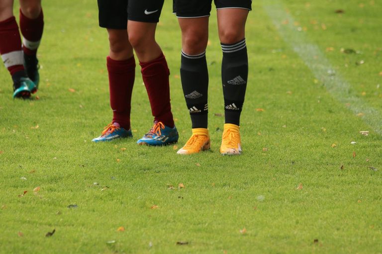 Zwei Fußballspieler, einer mit Adidas-Socken, stehen auf einem Feld mit blauen Schuhen, gelben Schuhen und roten Socken. Das Gras ist grün mit gefallenen Blättern.