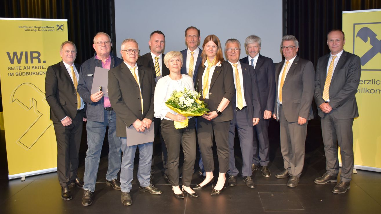 Eine Gruppe formell gekleideter Männer und Frauen steht zusammen auf der Bühne, zwei Frauen halten einen Blumenstrauß.