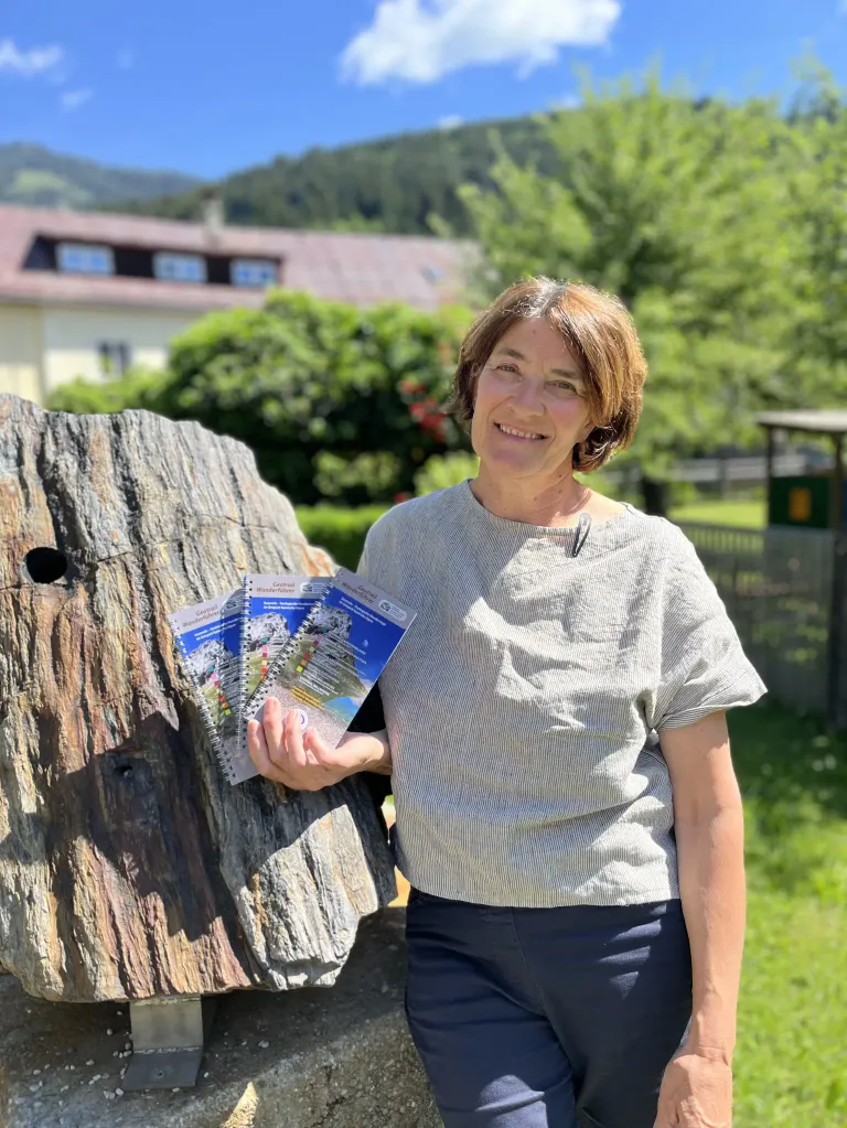 Eine Frau in einem grauen Oberteil und dunkler Hose hält einen Stapel Broschüren an einem Baumstamm. Sie lächelt und steht auf einem Rasen mit einem Haus und Bäumen im Hintergrund.