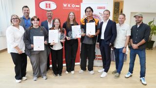 Sechs Personen stehen zusammen und halten Urkunden und Trophäen. Sie stehen vor einem roten Hintergrund mit Logos und Text. Die Gruppe besteht aus Männern und Frauen, alle in Freizeitkleidung.