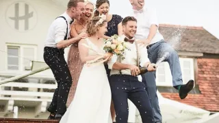 Eine Gruppe von Menschen, darunter ein Brautpaar, feiert auf einem Rasen vor einem Haus und pöppelt eine Champagnerflasche auf.