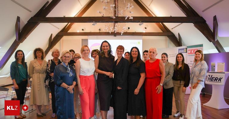Eine Gruppe von Frauen in formeller Kleidung posiert für ein Foto in einem Raum mit Holzbalken, einer Kristallleuchte und einer Projektionsleinwand.