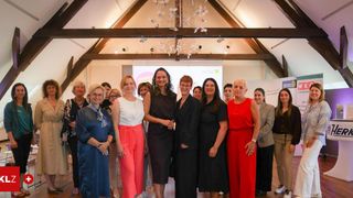 A group of women in formal attire pose for a photo in a room with wooden beams, a chandelier, and a projector screen.