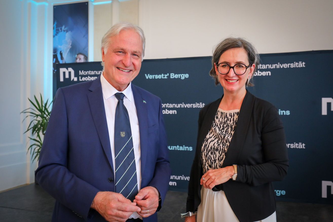 Ein Mann und eine Frau stehen lächelnd vor einer blauen Kulisse mit Logos.