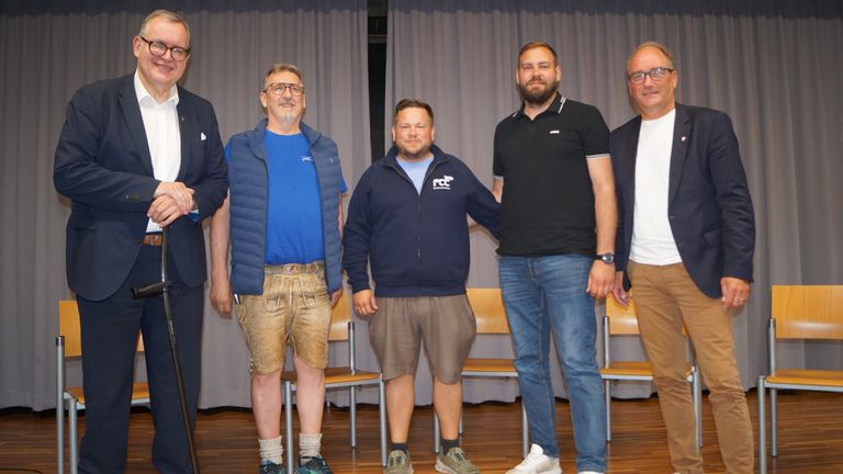 Fünf Männer stehen auf einer Bühne und lächeln für die Kamera. Zwei tragen Shorts, einer mit einem blauen Jackett und Logo, der andere in Braun. Der Mann auf der rechten Seite trägt ein schwarzes Hemd und Jeans.