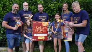 Eine Gruppe von Menschen steht hinter einem Schild für das Kelten Festival, wobei ein Mädchen eine Fackel und ein Schild hält und ein anderes Mädchen ein Poster hält. Sie tragen alle passende blaue T-Shirts.