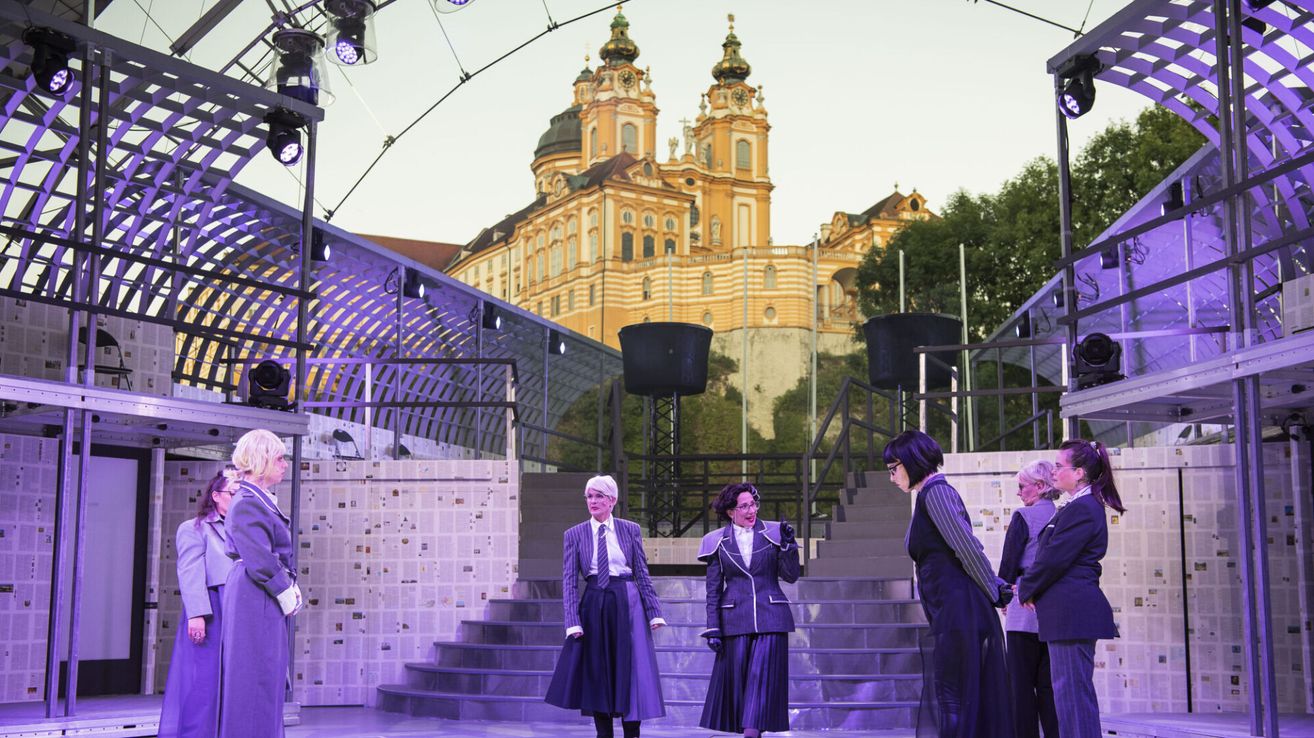 Vier Personen in formeller Kleidung stehen auf einer Bühne, mit einem großen Gebäude und einem Uhrturm im Hintergrund.