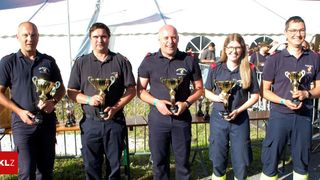 Drei Personen in Uniform posieren mit Trophäen in einem Zelt, einem Metallzaun und einem Rasen im Hintergrund.
