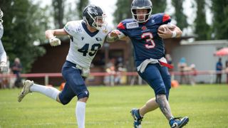 Zwei Footballspieler laufen auf einem Feld, einer in einem weißen Trikot und der andere in einem blauen Trikot. Der Spieler in Blau läuft mit einem Football.