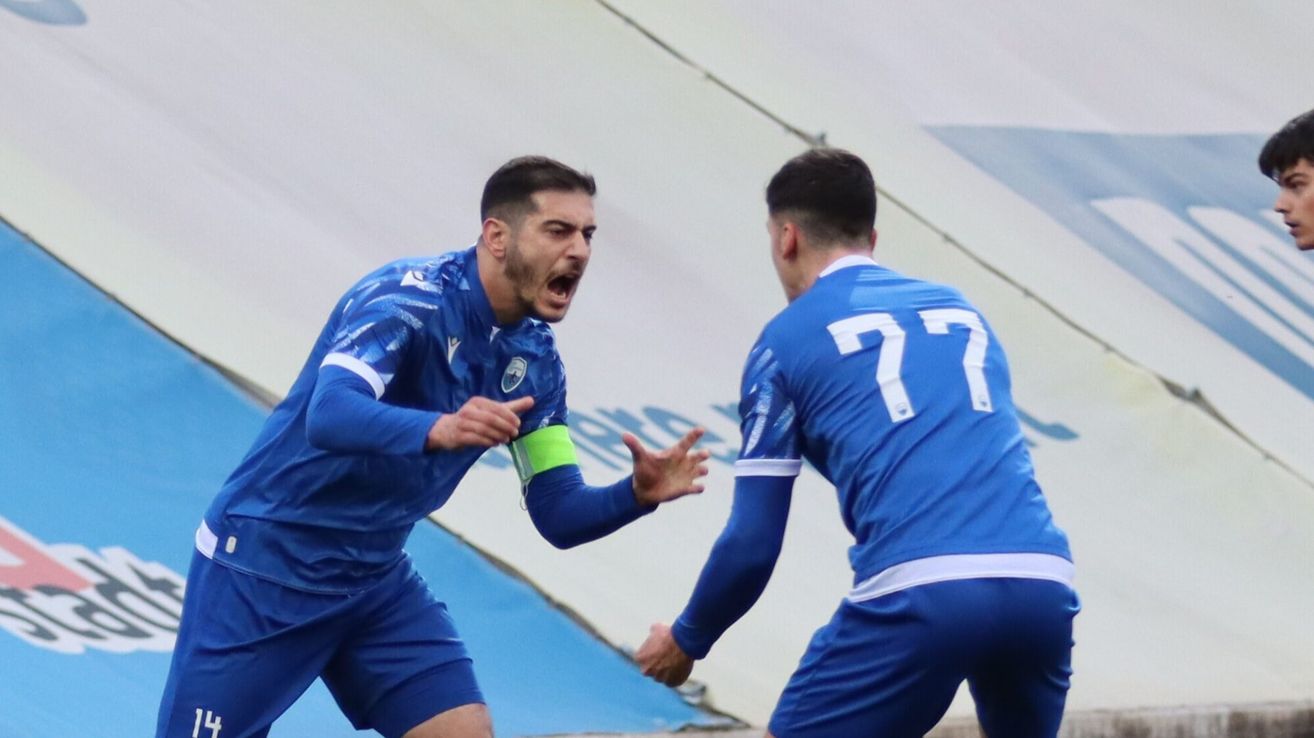 Zwei Fußballspieler in blauen Trikots. Ein Spieler schreit, während er läuft, und der andere schaut ihn an. Sie sind in der Nähe des Torbereichs.