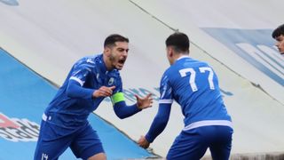 Zwei Fußballspieler in blauen Trikots. Ein Spieler schreit, während er läuft, und der andere schaut ihn an. Sie sind in der Nähe des Torbereichs.