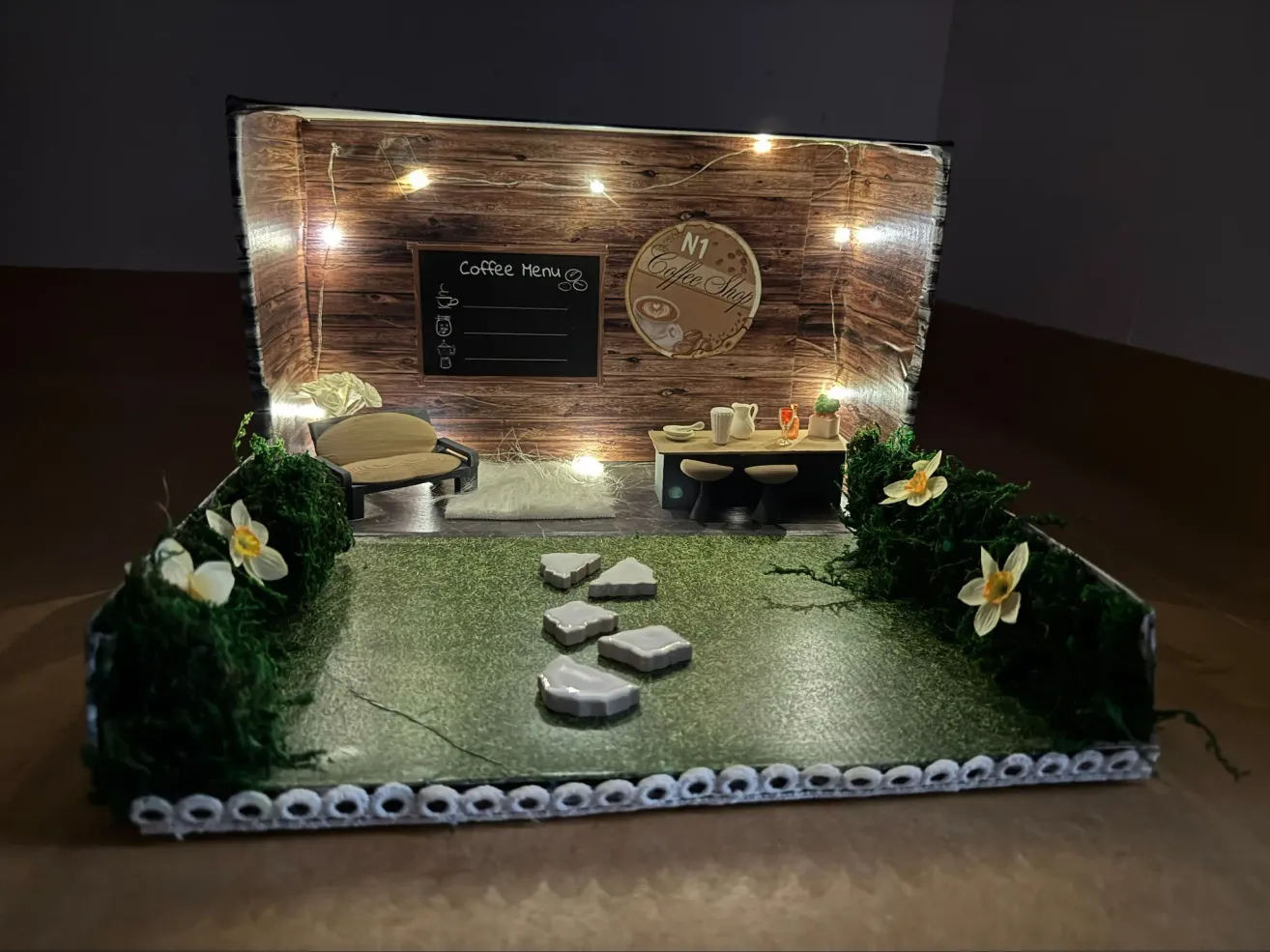 Eine Miniaturszene eines Cafés mit einer Holzwand und einer Menütafel. Der Bereich hat eine kleine Couch, einen Schreibtisch und dekorative Pflanzen. An der Wand sind Lampen befestigt.