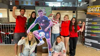 Eine Gruppe von Menschen in Sporttrikots und Brillen posiert mit einer großen Ausschnitt eines Fußballspielers mit der Nummer 14. Sie sind in einem hell erleuchteten Raum mit einer Leinwand, die ein Fußballspiel zeigt.