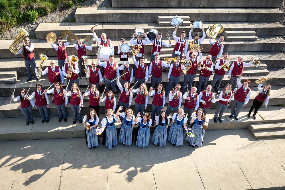 A large brass and percussion band poses on a set of stairs in traditional uniforms.