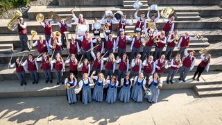 Ein großes Blech- und Perkussionsorchester posiert in traditioneller Uniform auf einer Treppe.
