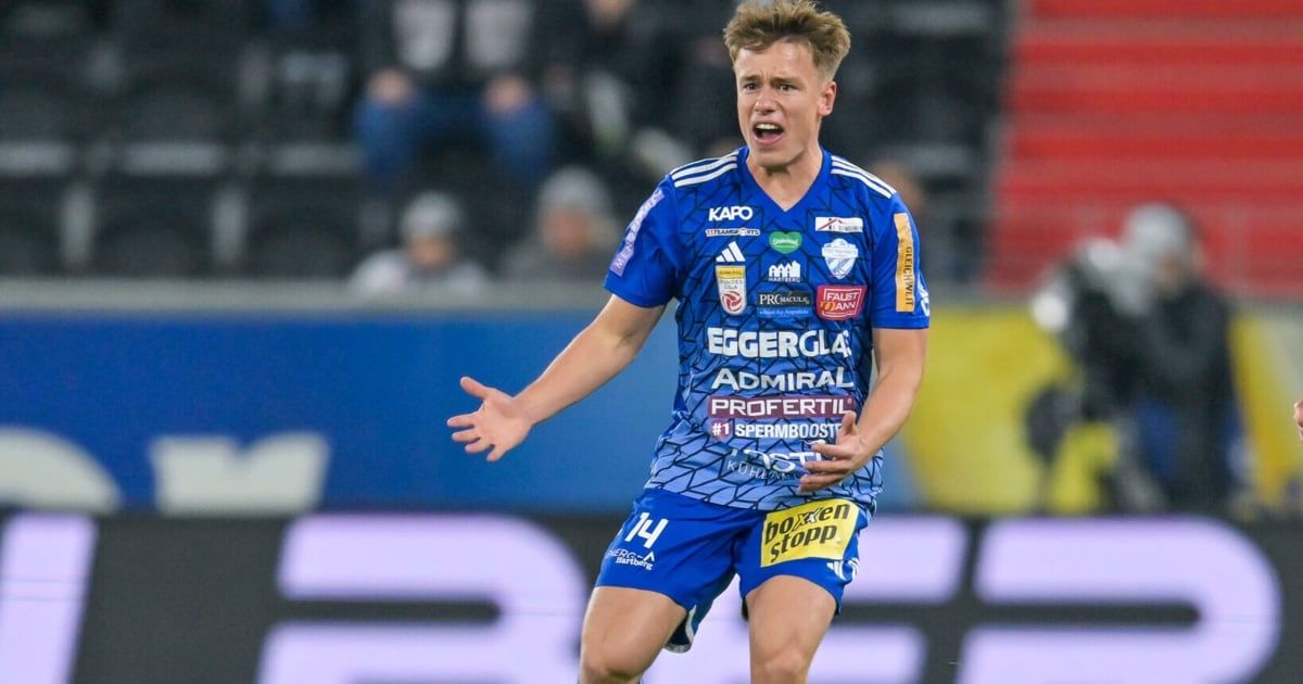 Ein Fußballspieler in einem blauen Trikot feiert ein Tor. Er trägt ein blau-weißes Trikot mit einem gelben Tag. Der Hintergrund ist verschwommen mit Menschen, die auf den Tribünen sitzen.
