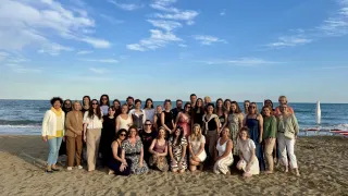 Eine große Gruppe von Menschen, wahrscheinlich ein Team, posiert für ein Foto auf einem Sandstrand unter einem klaren blauen Himmel mit einigen Wolken.