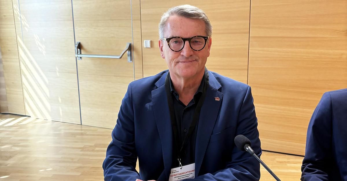 Ein Mann in einem blauen Anzug und mit einer Brille sitzt an einem Tisch mit einem Mikrofon vor ihm, wahrscheinlich an einer Pressekonferenz teilnehmend.