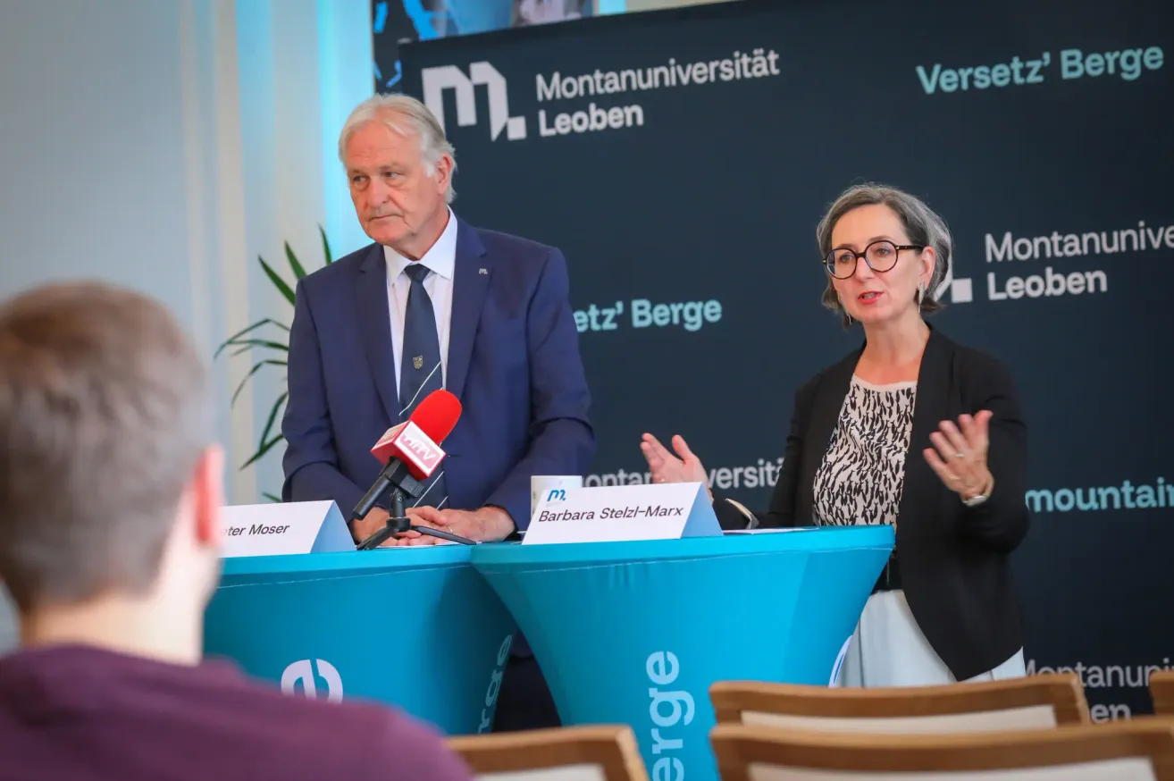 Zwei Personen, ein Mann und eine Frau, stehen an einem Podium mit Mikrofonen bei der Montanuniversität Leoben. Der Mann trägt einen Anzug mit Krawatte, die Frau eine Brille. Ein Namensschild trägt den Namen Barbara Stelzl-Marx. Dahinter ist ein schwarzer Hintergrund, auf dem das Logo der Montanuniversität Leoben steht.