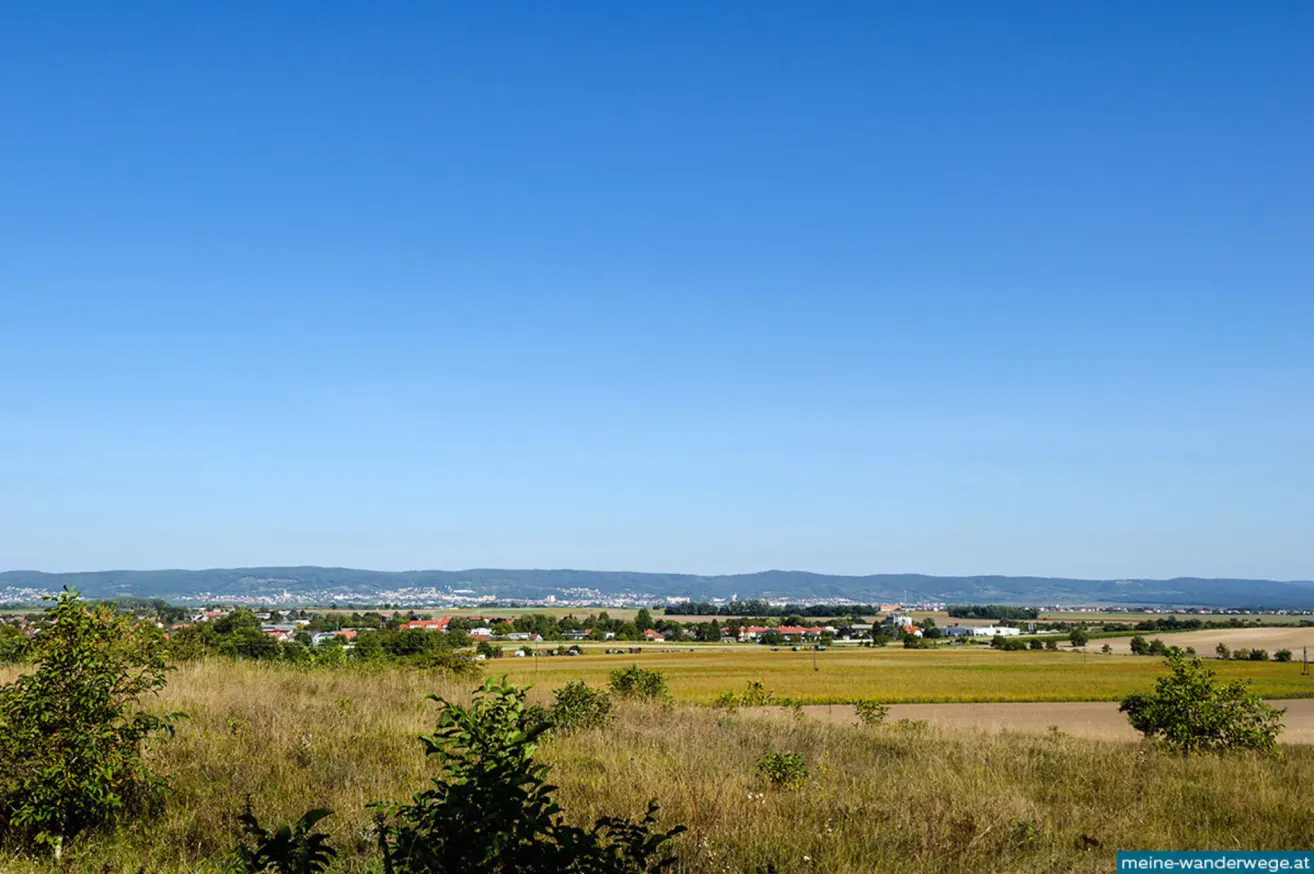 Ein weites Grasfeld mit einigen Bäumen im Vordergrund. In der Ferne ein Dorf mit Häusern und Gebäuden. Berge im Hintergrund unter einem klaren blauen Himmel.