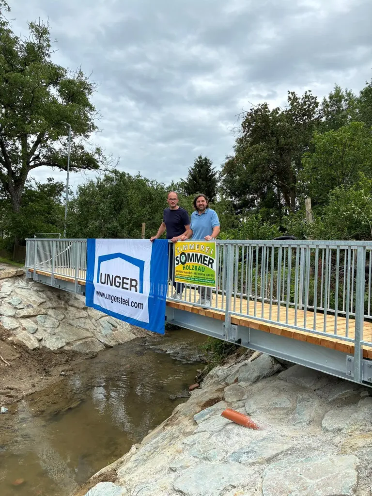 Zwei Männer stehen auf einer Brücke über einem Fluss. Einer hält ein blaues Banner mit 'UNGER' darauf. Der andere hält ein gelbes Schild mit 'SOMMER HOLZBAU'.