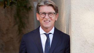 A man in a dark blue suit and tie, wearing glasses, smiles for the camera while leaning against a wall.