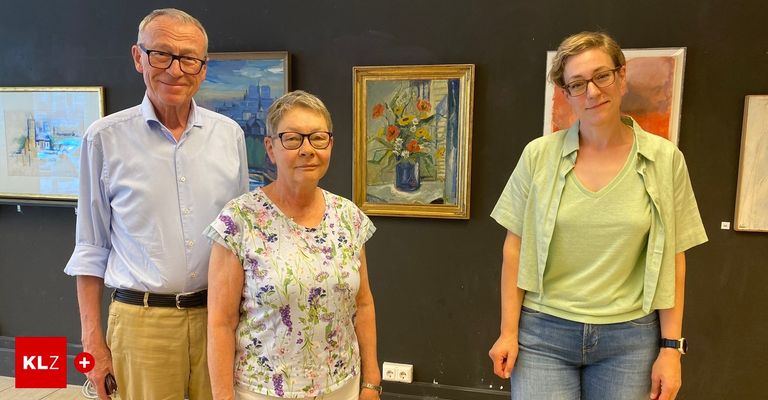 Drei Personen stehen neben einer schwarzen Wand mit Gemälden. Ein älterer Mann trägt eine Brille und einen Gürtel. Eine Frau neben ihm hat eine Brille und ein Blumenhemd. Eine junge Frau trägt eine Brille und ein grünes Shirt.