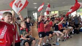 Gruppe junger Fußballfans in einem Raum, die Flaggen halten und Teamtrikots tragen. Der Raum ist mit Menschen gefüllt, einige sitzen und einige stehen, jubeln begeistert.