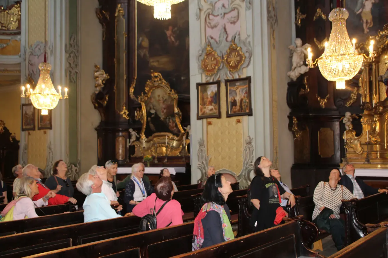 Eine Gruppe von Menschen sitzt in einer Kirche und schaut zu verzierten Decken auf. Die Wände sind mit Gemälden und Skulpturen verziert.