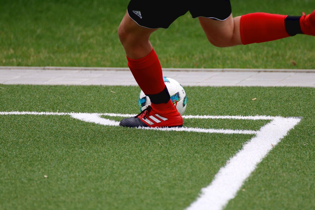 Ein Fußballspieler in roten Socken und Adidas-Schuhen tritt einen Ball auf Kunstrasen. Weiße Linien markieren die Feldgrenzen.