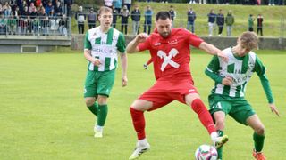 Zwei Fußballspieler in grünen und roten Trikots spielen auf einem Rasenfeld. Der Spieler in Rot ist dabei, den Ball zu schießen, während der Spieler in Grün ihn ansieht.