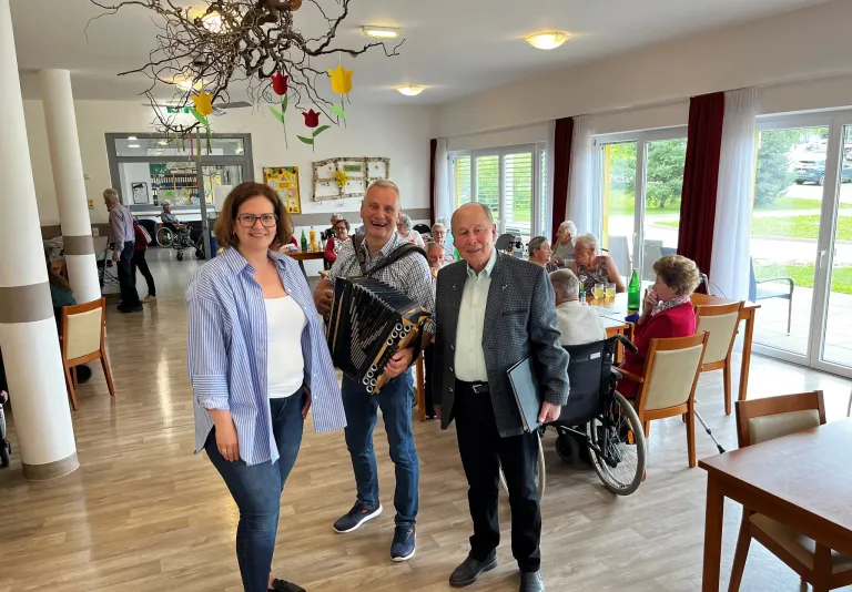 Drei Menschen stehen in einem Esszimmer mit älteren Personen, die an Tischen sitzen und stehen, während ein Mann Akkordeon spielt.
