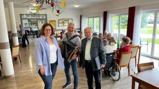 Drei Menschen stehen in einem Esszimmer mit älteren Personen, die an Tischen sitzen und stehen, während ein Mann Akkordeon spielt.