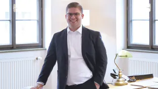 A man in a business suit stands smiling in an office with a desk lamp in the background.