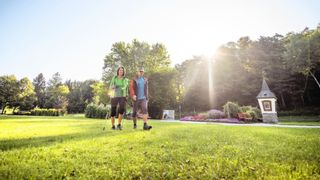 Zwei Personen, eine Frau und ein Mann, gehen mit Stöcken auf einem grünen Rasen. Dahinter sind Bäume, Blumen, Bänke und eine Straßenlampe. Die Sonne scheint hell.