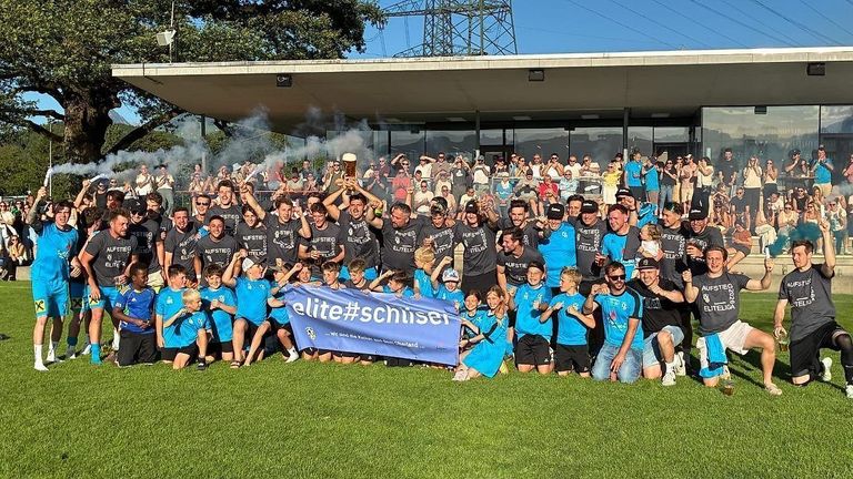 Eine Sportmannschaft feiert auf einem Feld mit einem großen Banner. Sie tragen blaue Trikots und einige halten Pokale. Die Menge hinter ihnen schaut zu und jubelt.
